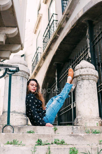 Young casual woman sitting on old stairway lifting legs on stone pillar and looking at camera — Stock Photo