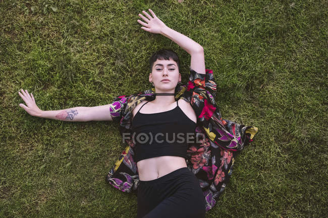 Зверху молода приваблива стильна жінка в барвистій сорочці лежить на зеленій траві, простягаючи руки. — стокове фото