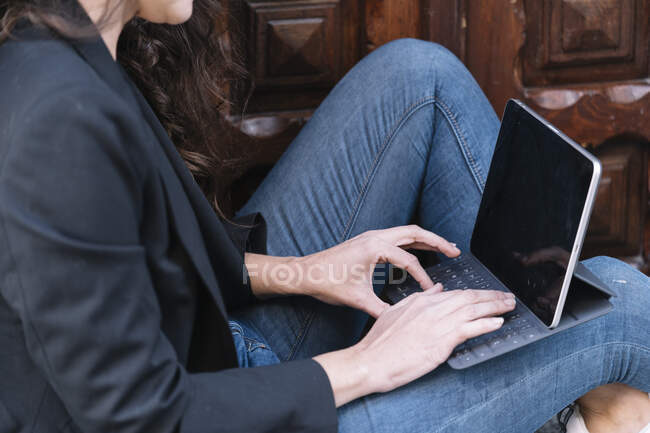 Вид збоку на невпізнавану жінку з використанням цифрового планшета, розслабляючись на порозі старих дерев'яних дверей — стокове фото