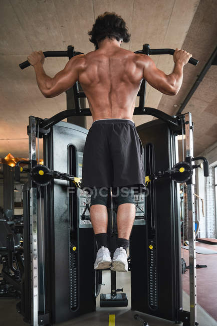 Rückenansicht eines hemdslosen Mannes, der Klimmzüge an einem Trainingsgerät macht, während er im Fitnessstudio trainiert — Stockfoto