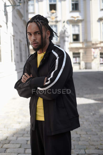 Молодой этнический человек с плетеными волосами в черном спортивном костюме смотрит в камеру на городском фоне — стоковое фото