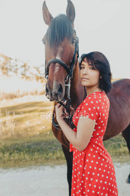 Mujer joven en vestido rojo largo whit caballo, mientras que de pie en el patio del rancho en el día soleado en el campo - foto de stock