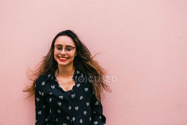 Encantadora joven con gafas y camisa sonriendo y mirando a la cámara contra la pared rosa - foto de stock