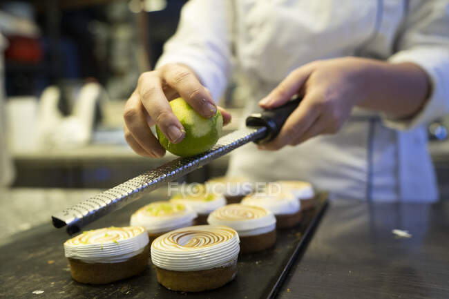 Crop confectioner in white uniform decorating delicious baked cakes with zest of lime in kitchen - foto de stock