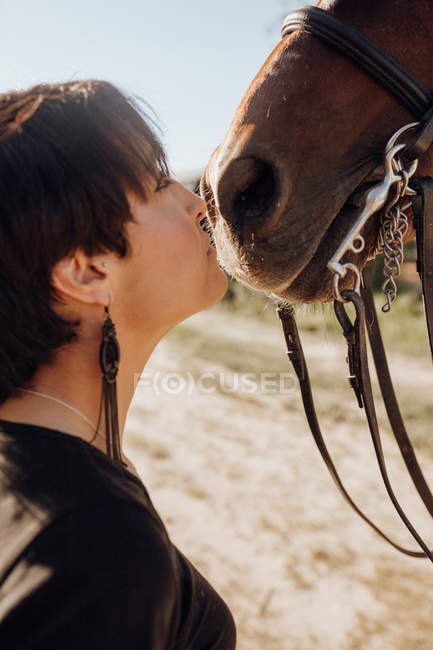 Молодая женщина с закрытыми глазами целует лошадь, проводя время на ранчо в солнечный день — стоковое фото