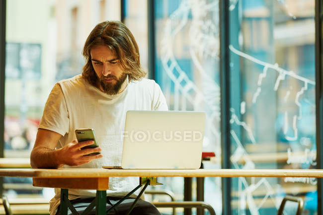 Joven hombre guapo barbudo sentado en la cafetería y trabajando sosteniendo el teléfono y el ordenador portátil en la mesa - foto de stock