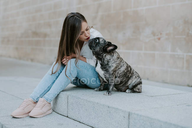 Casual young woman sitting on concrete pavement and kissing adorable bulldog on street — Stock Photo