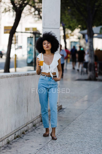 Веселий модний афроамериканець жінка п'є апельсиновий сік і дивлячись на камеру на міському фоні — стокове фото