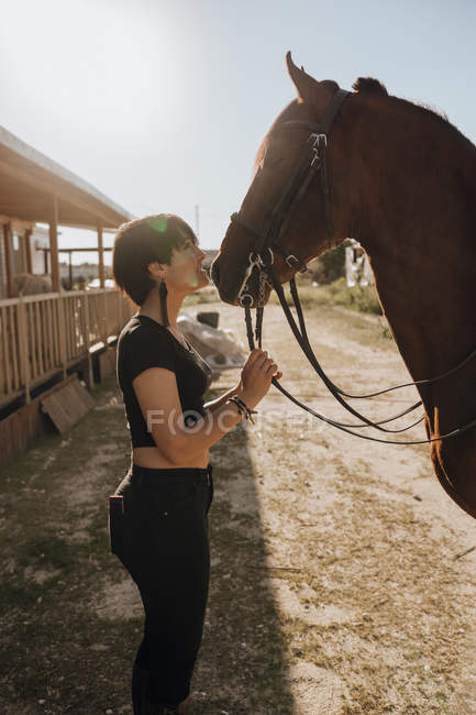 Mujer joven con los ojos cerrados besando caballo mientras pasa tiempo en el rancho en el día soleado - foto de stock
