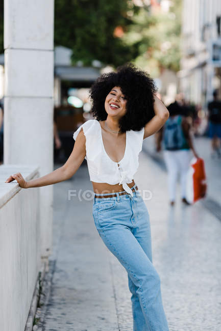 Cheerful African American woman in jeans and crop top relaxing on stone railing and looking at camera outdoors — Stock Photo