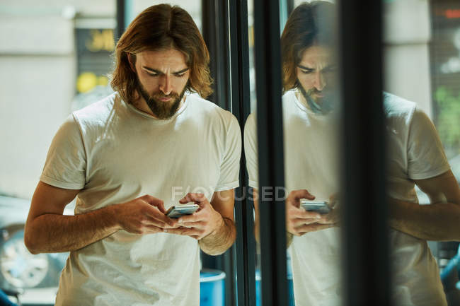Молодой бородатый красивый мужчина держит мобильный телефон и обменивается сообщениями, опираясь на зеркальную поверхность — стоковое фото