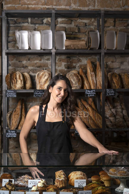 Красива жінка в чорному фартусі продає французьку багету в пекарні. — стокове фото