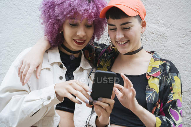 Young attractive women with black and pink hair in bright clothes laughing and embracing while watching on mobile phone on grey background — Stock Photo