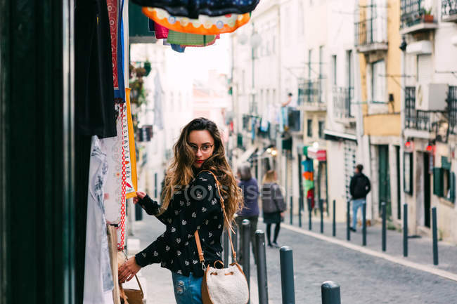 Молодая женщина смотрит на красочную одежду для продажи стоя на киоске на улице старого города — стоковое фото