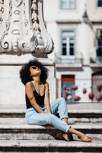 Ethnic woman in jeans and tank top relaxing and sunbathing on stone stairs against urban background — Stock Photo