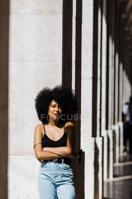 Young ethnic woman in jeans and tank top leaning on wall outdoors — Stock Photo