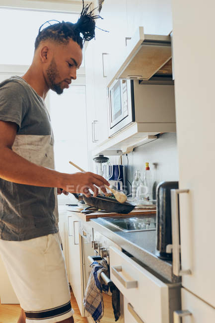 African American braided man standing in kitchen with frying pan and scapula — Stock Photo