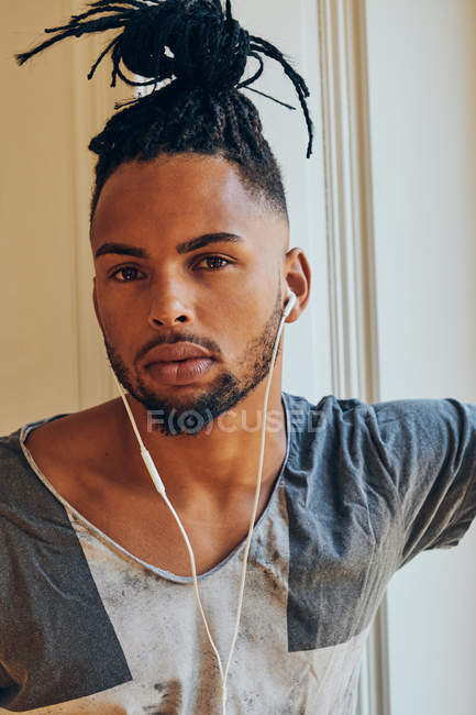 Serious ethnic man with creative hairstyle listening to music with earphones and looking at camera — Stock Photo