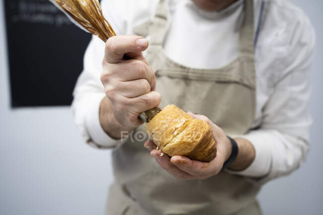 Кукурудзяна людина в фартусі та білій однорідній начинці свіжий круасан з шоколадним кремом у приміщенні — стокове фото