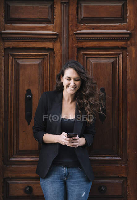 Красивая веселая женщина с помощью мобильного телефона, расслабляясь опираясь на старую деревянную дверь — стоковое фото