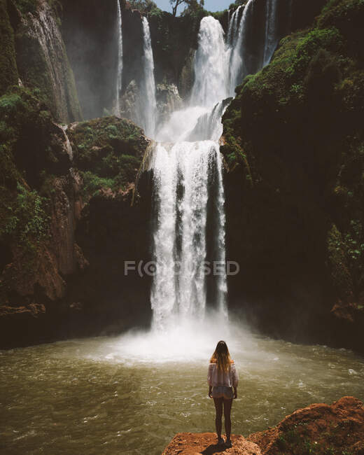 Back view of woman standing in desert terrain with powerful majestic waterfall on green cliffs, Morocco — Stock Photo