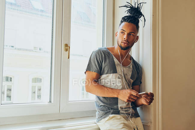Молодий афроамериканець людина з творчою зачіскою спираючись на підвіконня в домашніх умовах за допомогою мобільного телефону і прослуховування музики з навушниками — стокове фото