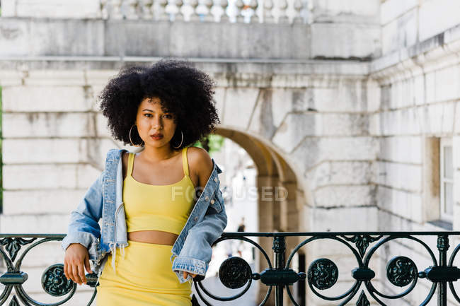 Mulher afro-americana em terno amarelo e jaqueta de ganga de pé e olhando para a câmera no fundo urbano — Fotografia de Stock