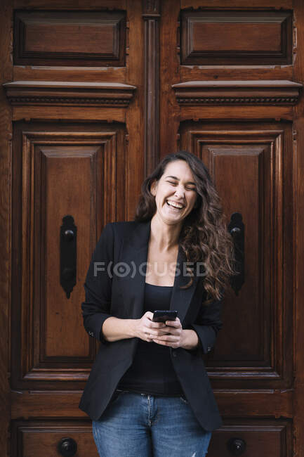 Hermosa mujer alegre usando el teléfono móvil mientras se relaja apoyado en la vieja puerta de madera - foto de stock
