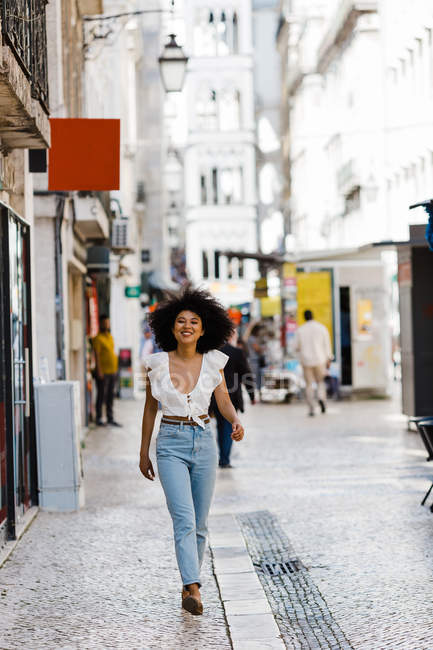 Happy ethnic woman in trendy outfit walking and looking at camera on summer day — Stock Photo