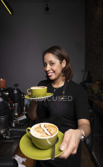 Smiling female barista giving green coffee cups with latte art and looking at camera in cafe — Stock Photo