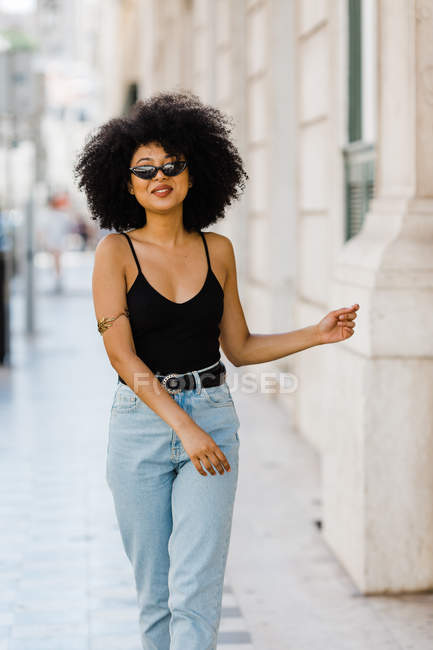 Jeune femme ethnique en jeans et débardeur marchant et souriant à la caméra à l'extérieur — Photo de stock