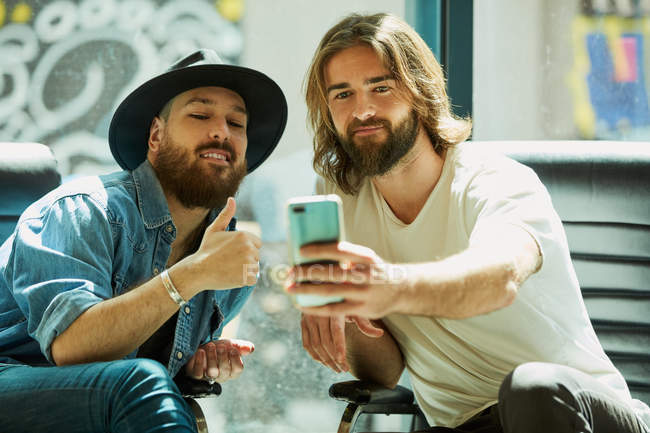 Handsome man in black hat sitting and enjoying process of taking selfie on mobile phone with friend in cafe — Stock Photo