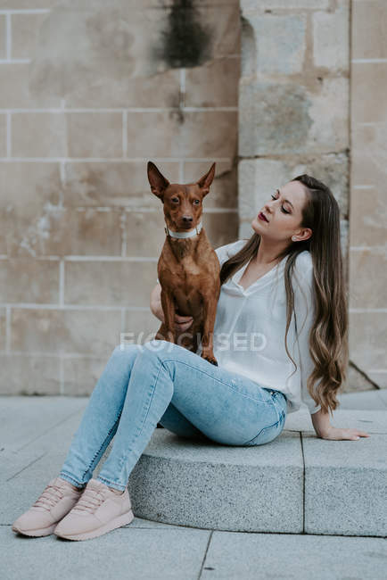 Mulher casual com cão de caça de joelhos sentado em passo concreto na rua e olhando para longe — Fotografia de Stock