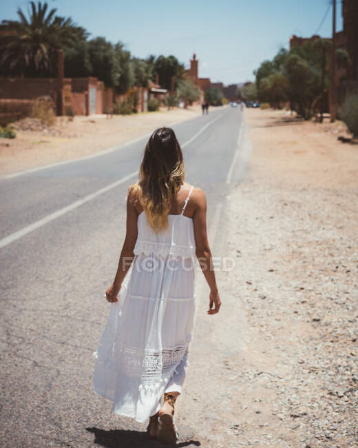 Обратный вид женщины в белом летнем платье во время прогулки по улице пустыни Марокко — стоковое фото