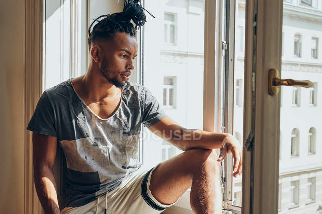 Pensiero uomo afroamericano con trecce seduto a casa sul davanzale della finestra — Foto stock