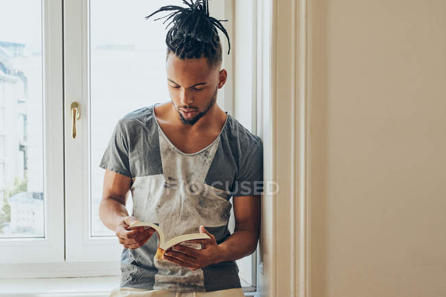 Junger afrikanisch-amerikanischer Mann lehnt auf Fensterbank und liest Buch — Stockfoto