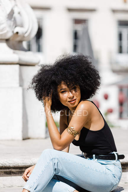 Portrait of ethnic woman in jeans and tank top relaxing and sunbathing  against urban background — curly, hairstyle - Stock Photo | #285629690
