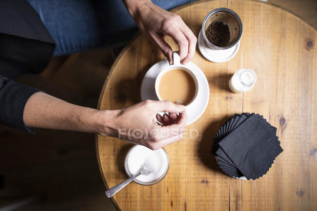 De arriba cosechan las manos de la hembra sentada a la mesa de madera y revolviendo el té caliente con la leche y el azúcar - foto de stock