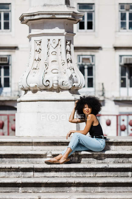 Donna etnica in jeans e canotta relax e prendere il sole su scale in pietra sullo sfondo urbano — Foto stock