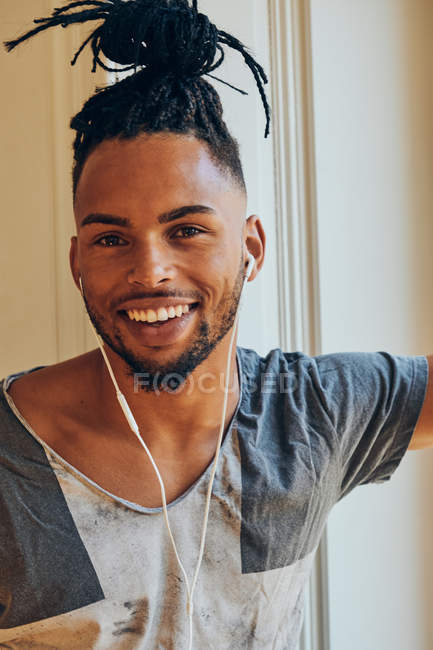 Homme afro-américain souriant avec des tresses écouter de la musique avec des écouteurs à la maison sur fond de fenêtre — Photo de stock