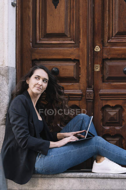 Vista lateral da mulher bonita usando tablet digital enquanto relaxa no limiar da velha porta de madeira — Fotografia de Stock