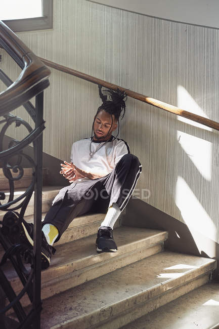 Beau Afro-Américain avec des cheveux tressés assis sur les escaliers et regardant les mains — Photo de stock