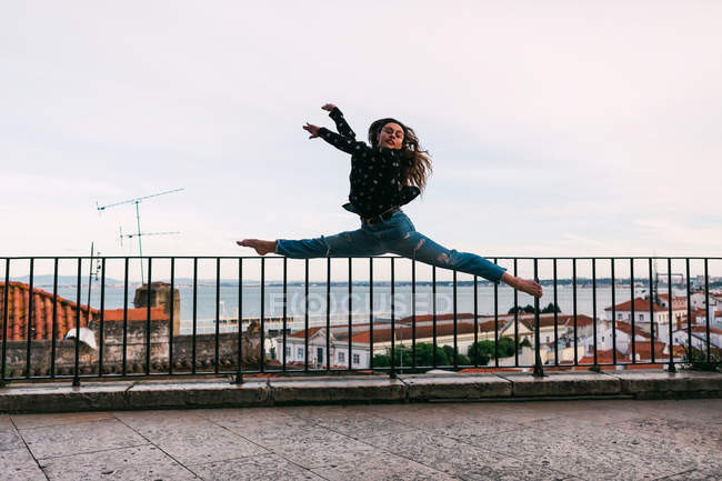 Casual joven mujer volando en splits mientras baila sobre fondo de la ciudad costera - foto de stock