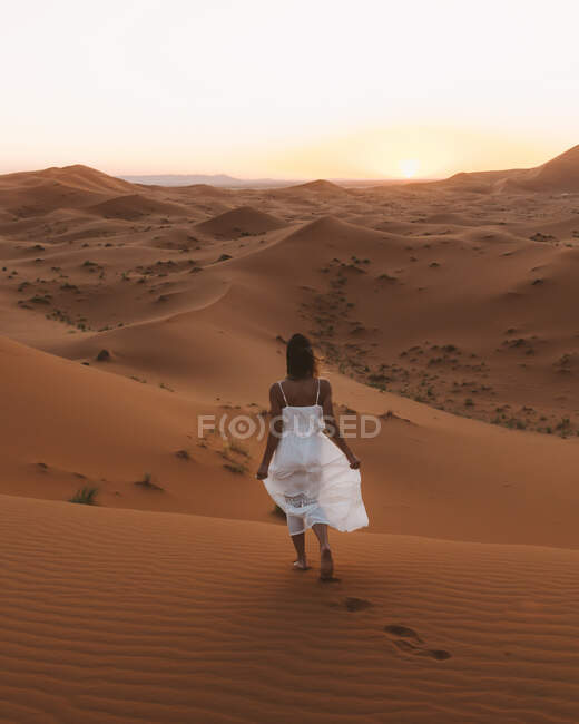 Vista posteriore delle donne scalze in abito bianco estivo che camminano su dune sabbiose di deserto infinito al tramonto, Marocco — Foto stock