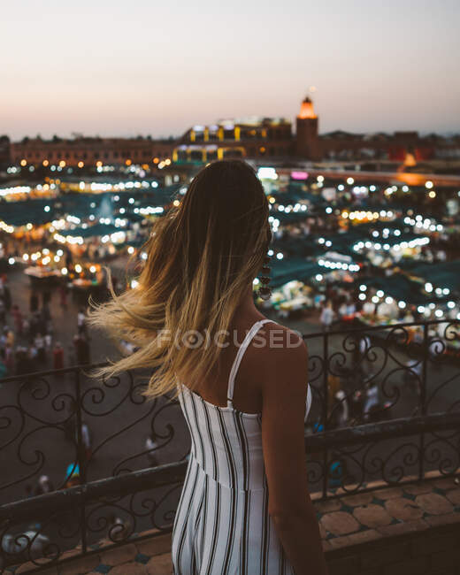 Rückansicht einer trendigen Frau mit fliegendem Haar, die auf dem Balkon vor hellem Licht der marokkanischen Stadt in der Dämmerung steht — Stockfoto