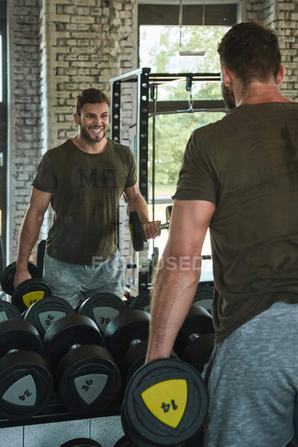 Strong man exercising with dumbbells in gym in front of mirror — Stock Photo