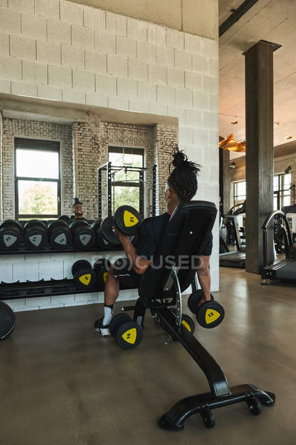 Strong black man exercising with dumbbells in gym in front of mirror — Stock Photo