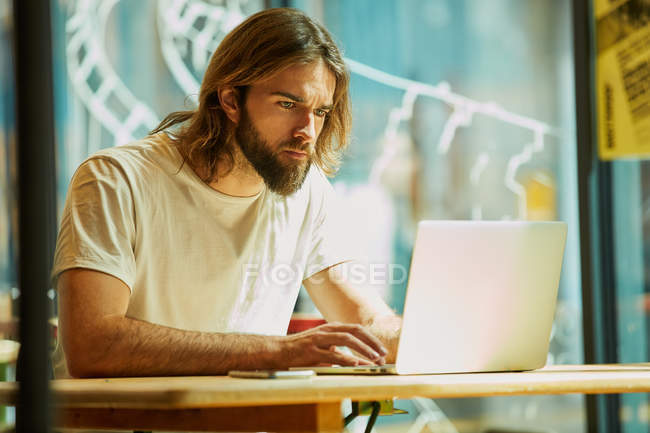 Joven hombre guapo barbudo sentado en la cafetería exterior y trabajando con el ordenador portátil en la mesa - foto de stock