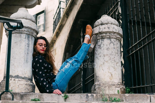 Joven mujer casual sentado en la vieja escalera levantamiento de piernas en pilar de piedra - foto de stock