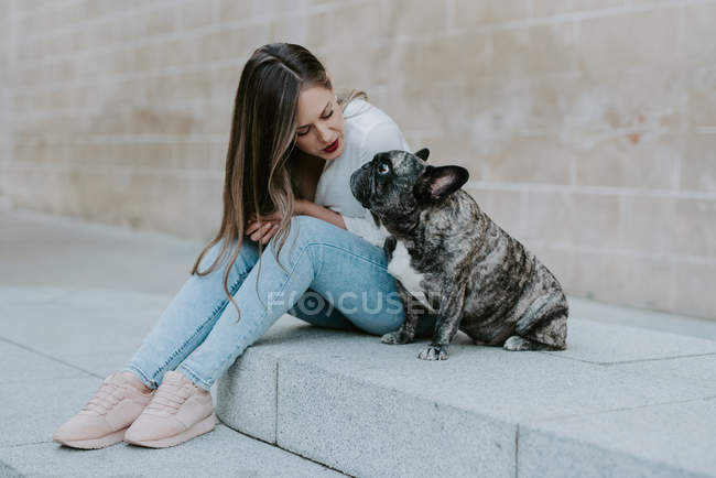 Jovem mulher sentada no pavimento de concreto com buldogue adorável, olhando uns para os outros — Fotografia de Stock